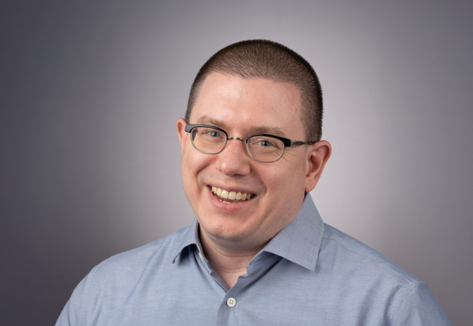 A color photo of Michael Clarkson, a smiling man with a buzzed head, glasses and a gray shirt in front of a gray background