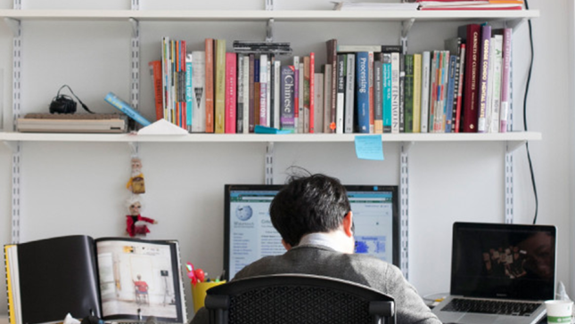 Graduate student working at a desk with books, monitors, and a laptop.