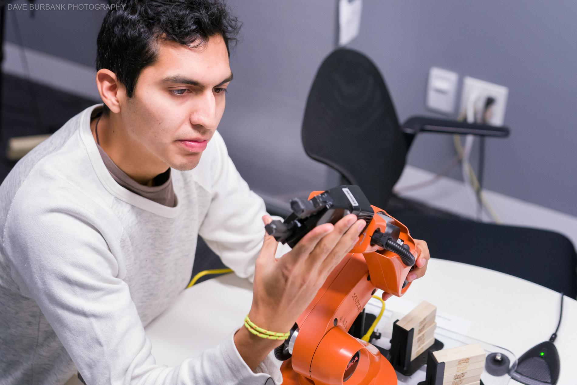 a male student with tan skin and dark black hair touches an orange robot